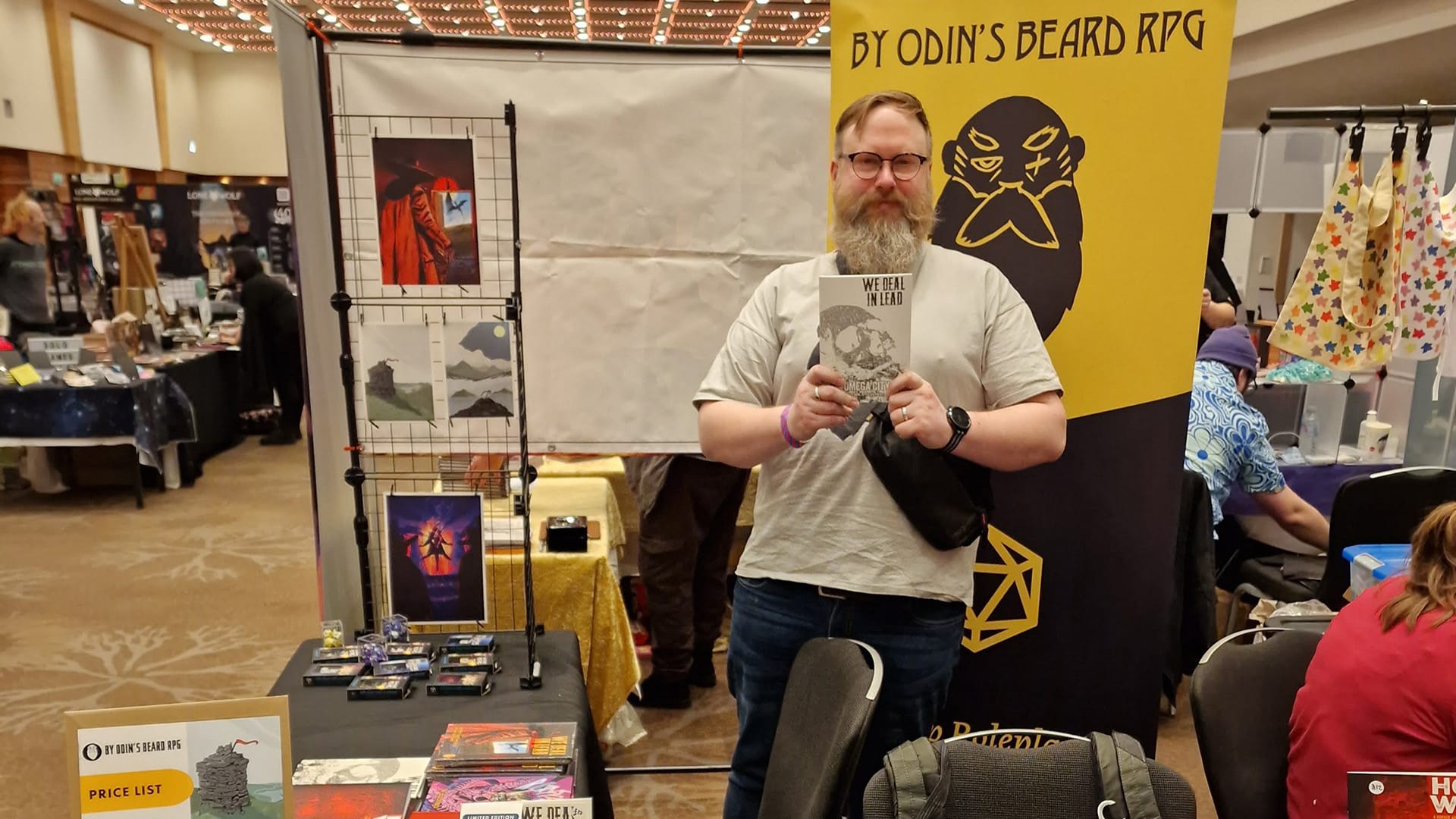 Me (Colin), a white man with a beard and glasses, holds a copy of We Deal in Lead Omega City behind a convention stand