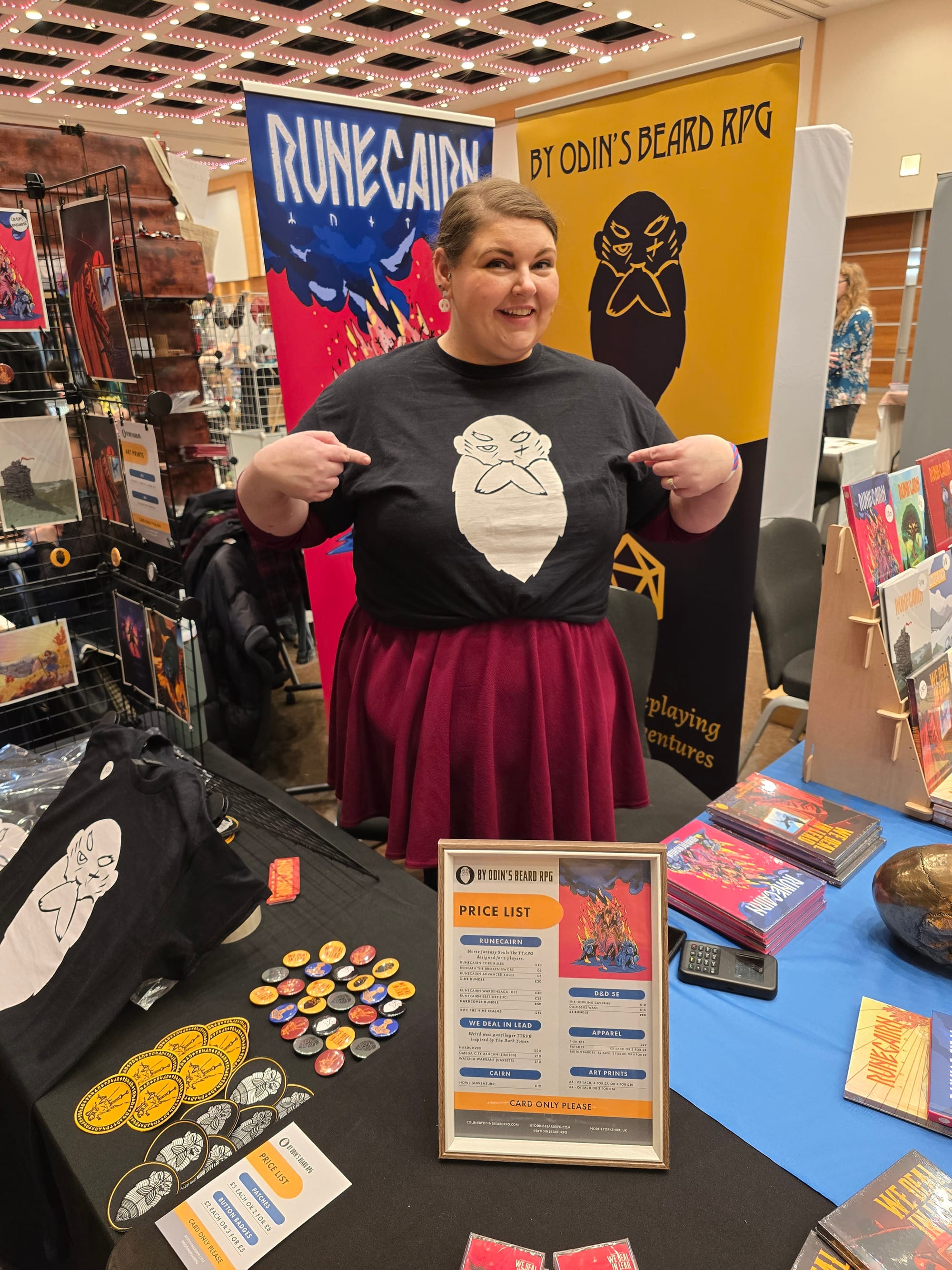 A white woman with brown hair, smiling while pointing at her black t-shirt.