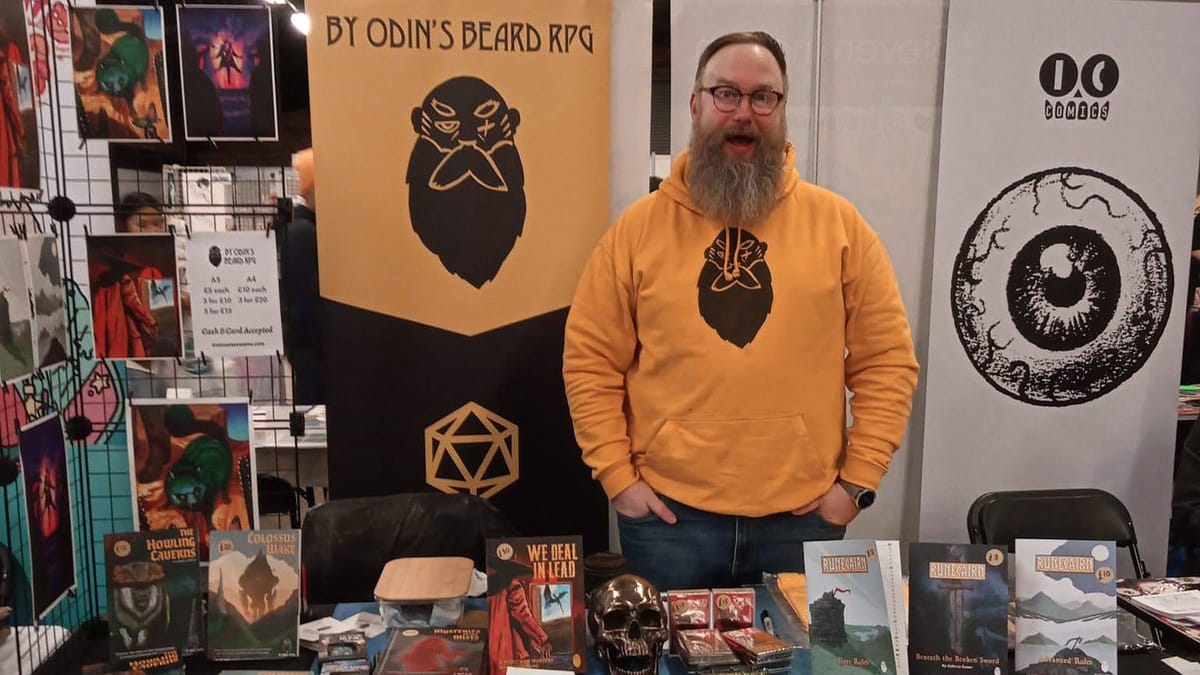 A white man with a beard and glasses, wearing a yellow By Odin's Beard RPG hoodie and looking happ